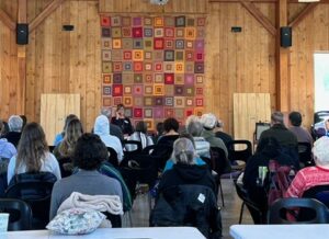 Practitioners sitting in a retreat center, facing away from the view.  A teacher sits in front, with a colorful quilted wall hanging against a wooden wall in the background.